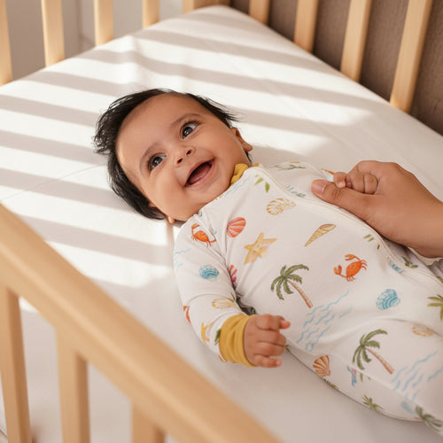 Baby lying in a crib wearing a swaddle with beach-themed print, held by an adult hand.