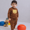 Child wearing a brown outfit with a bear design, standing in front of colorful toys on a white floor.