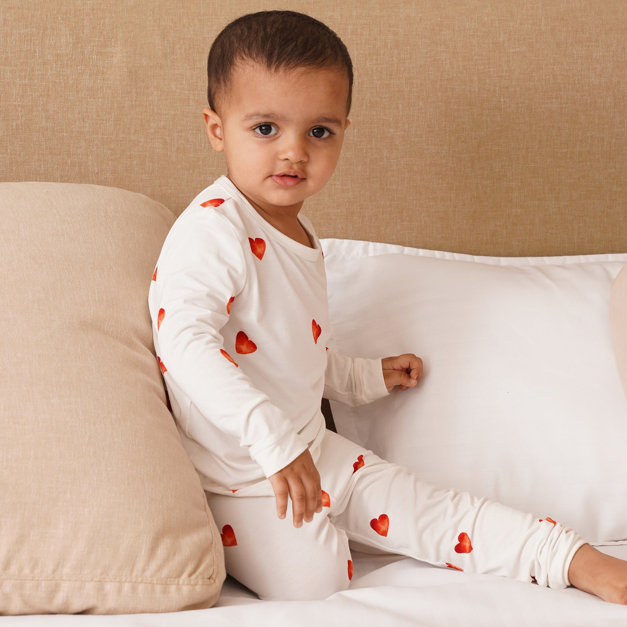Child wearing a bamboo white outfit with red heart patterns sitting on a bed.