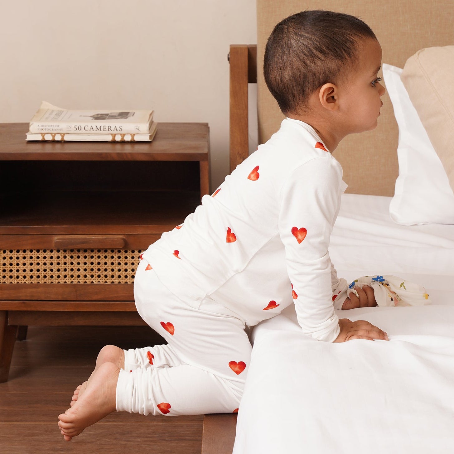 Child in bamboo white pajamas with red hearts sitting on a bed next to a wooden nightstand.
