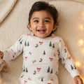 Child wearing a white long-sleeve shirt with Christmas tree and bow pattern, lying on a soft surface with lights in the background.