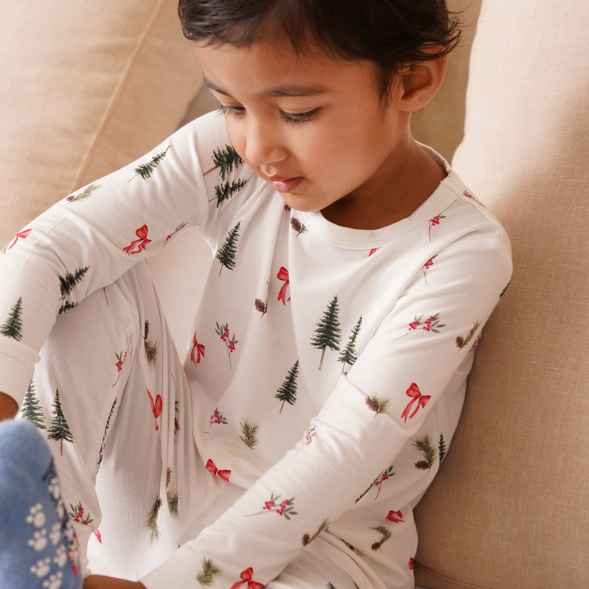 Child wearing a white bamboo long-sleeve shirt with Christmas tree and bow pattern, sitting on a beige couch.