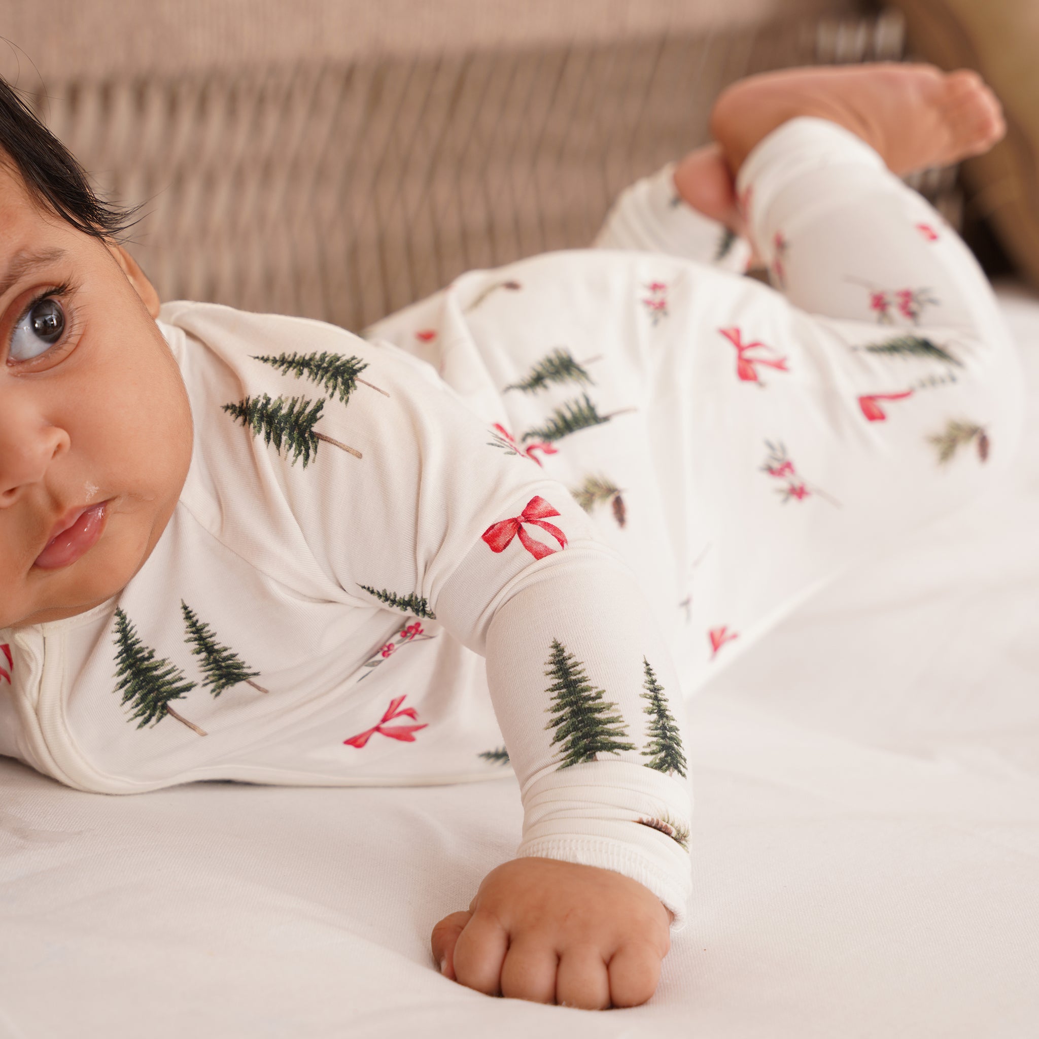 Baby wearing a white bamboo onesie with Christmas tree and bow pattern on a bed.