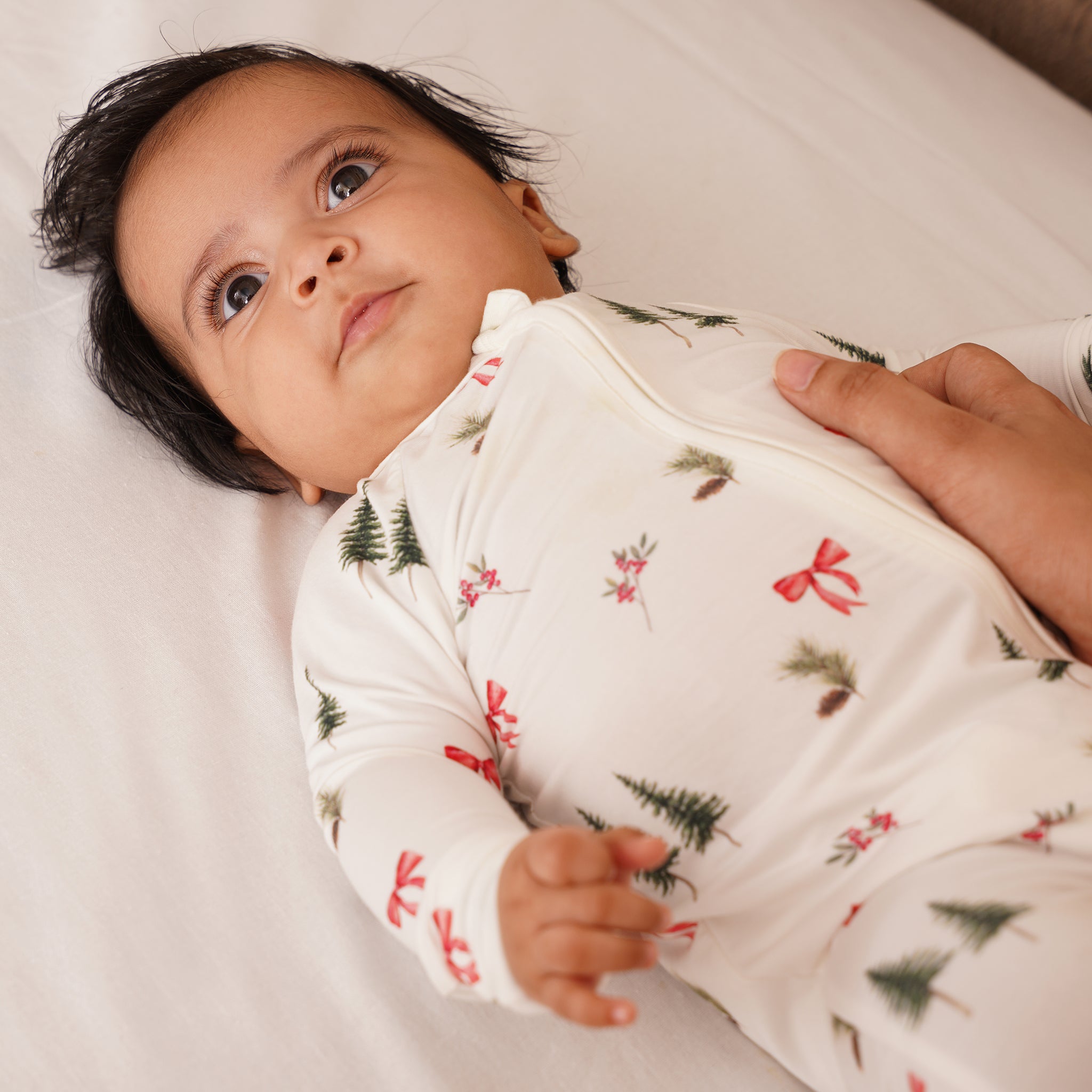 Baby wearing a white bamboo onesie with green tree patterns, lying on a white surface.