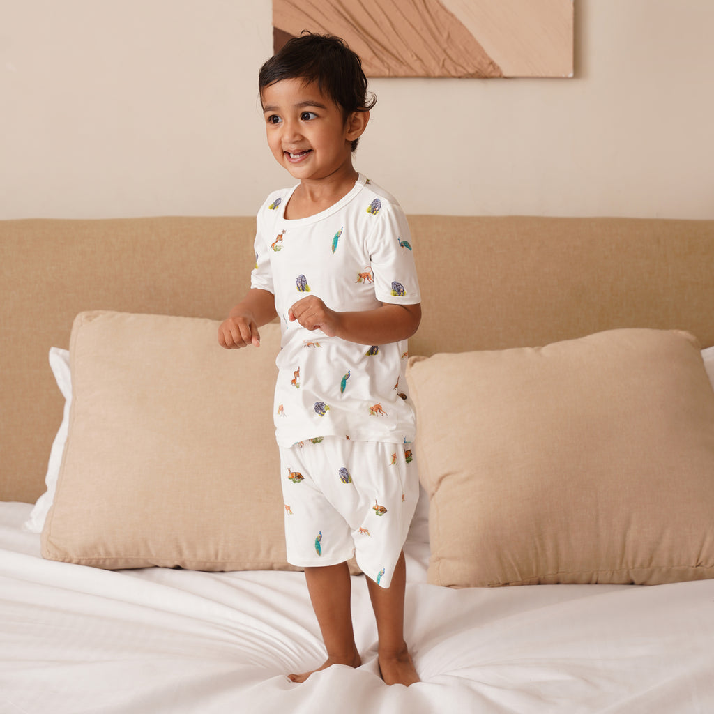 Child in a bamboo white outfit with animal prints standing on a bed with beige pillows and a neutral headboard.