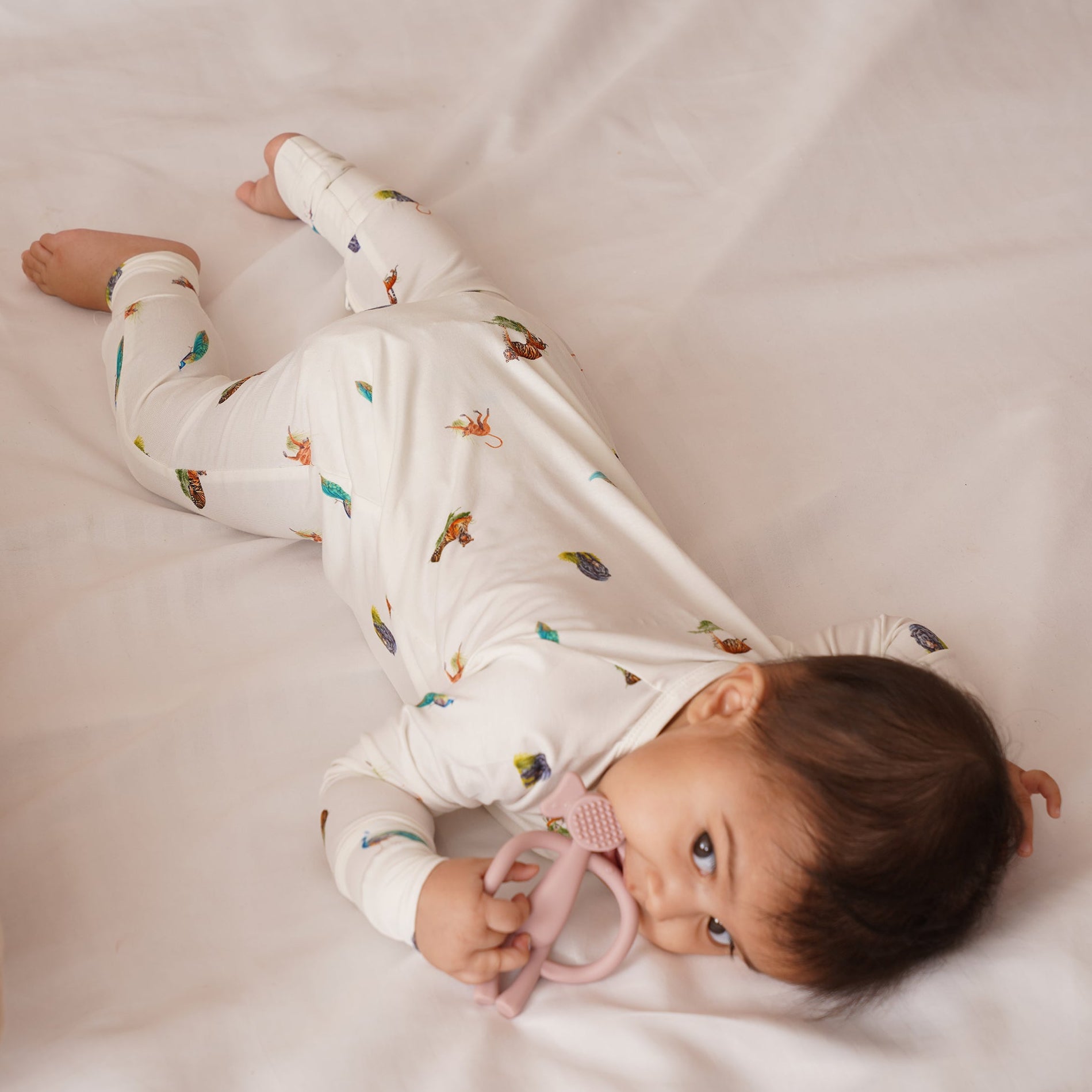 Baby lying on a white blanket holding pink teether, wearing bamboo onesie