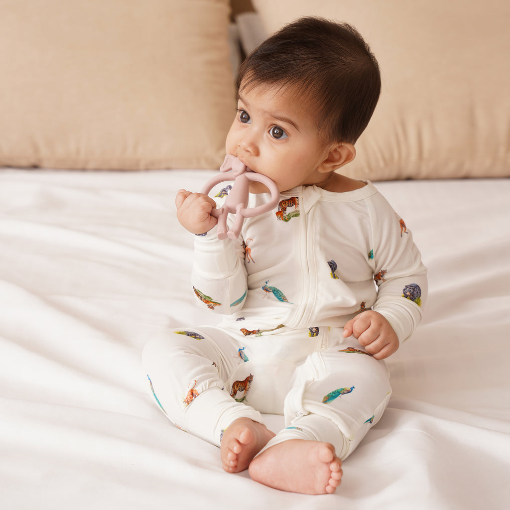 Baby wearing a white  bamboo onesie with colorful patterns, sitting on a bed holding a pink teething ring.
