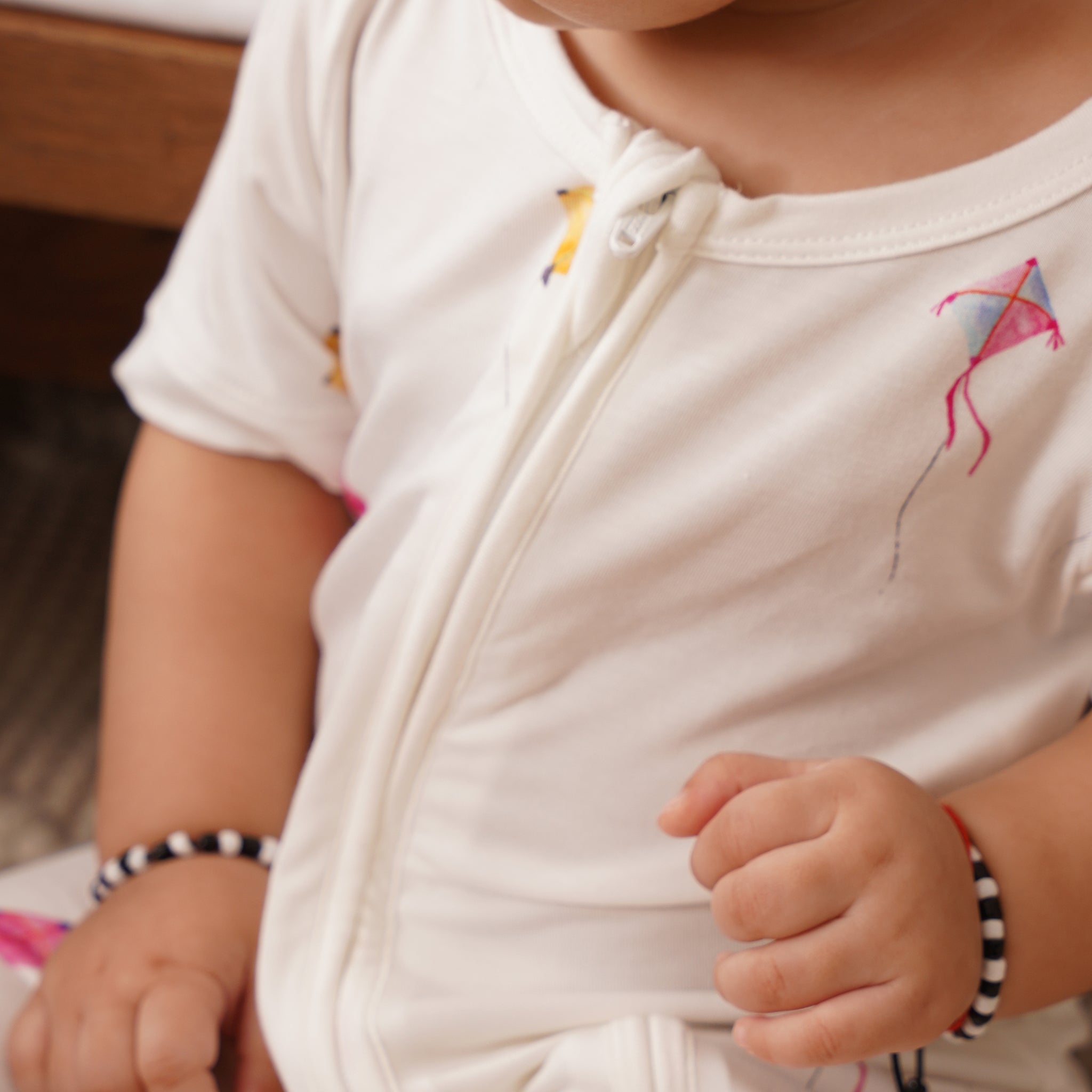 Child wearing a white bamboo outfit with kite designs, sitting on a wooden floor.