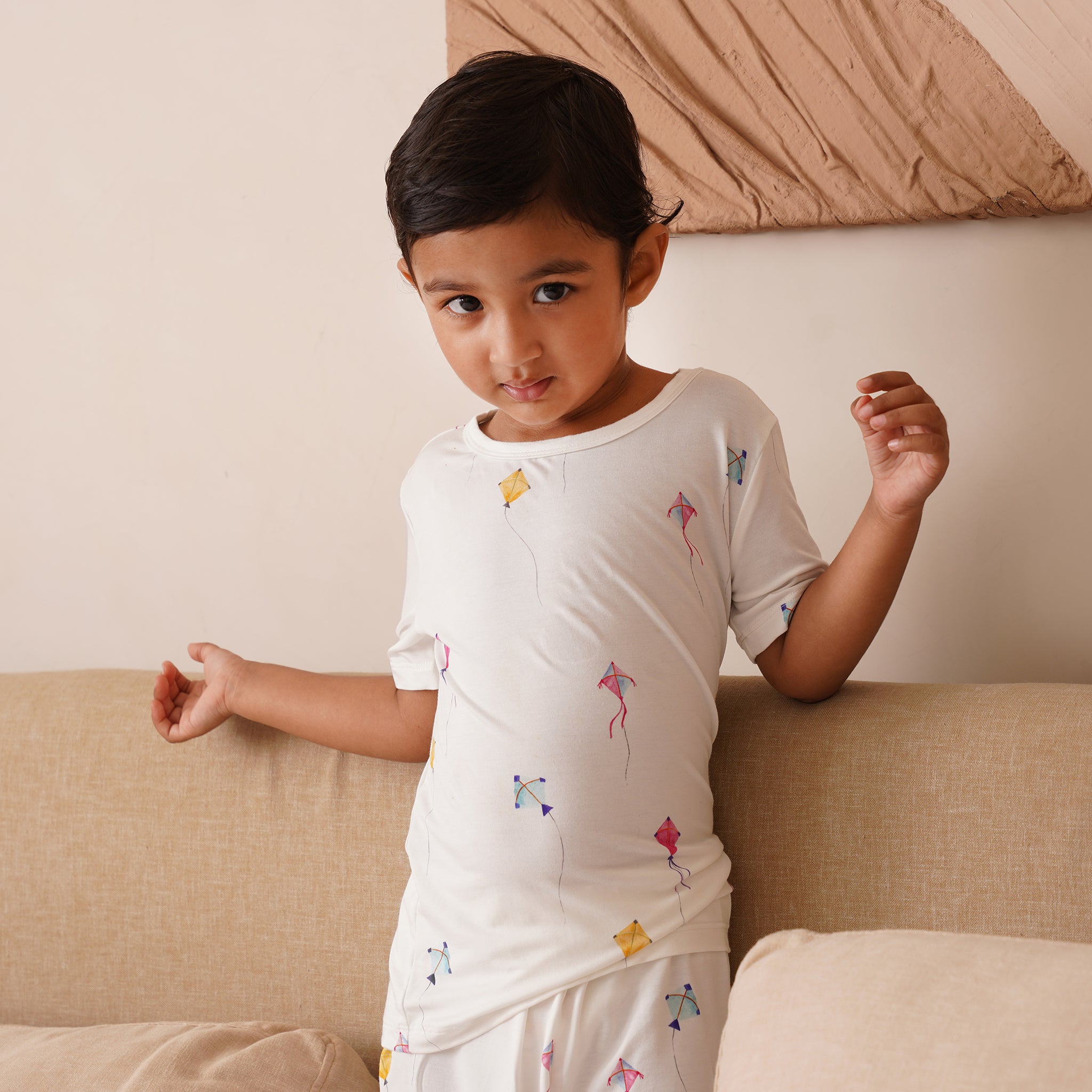 Child wearing a white bamboo outfit with colorful designs, standing on a beige couch.