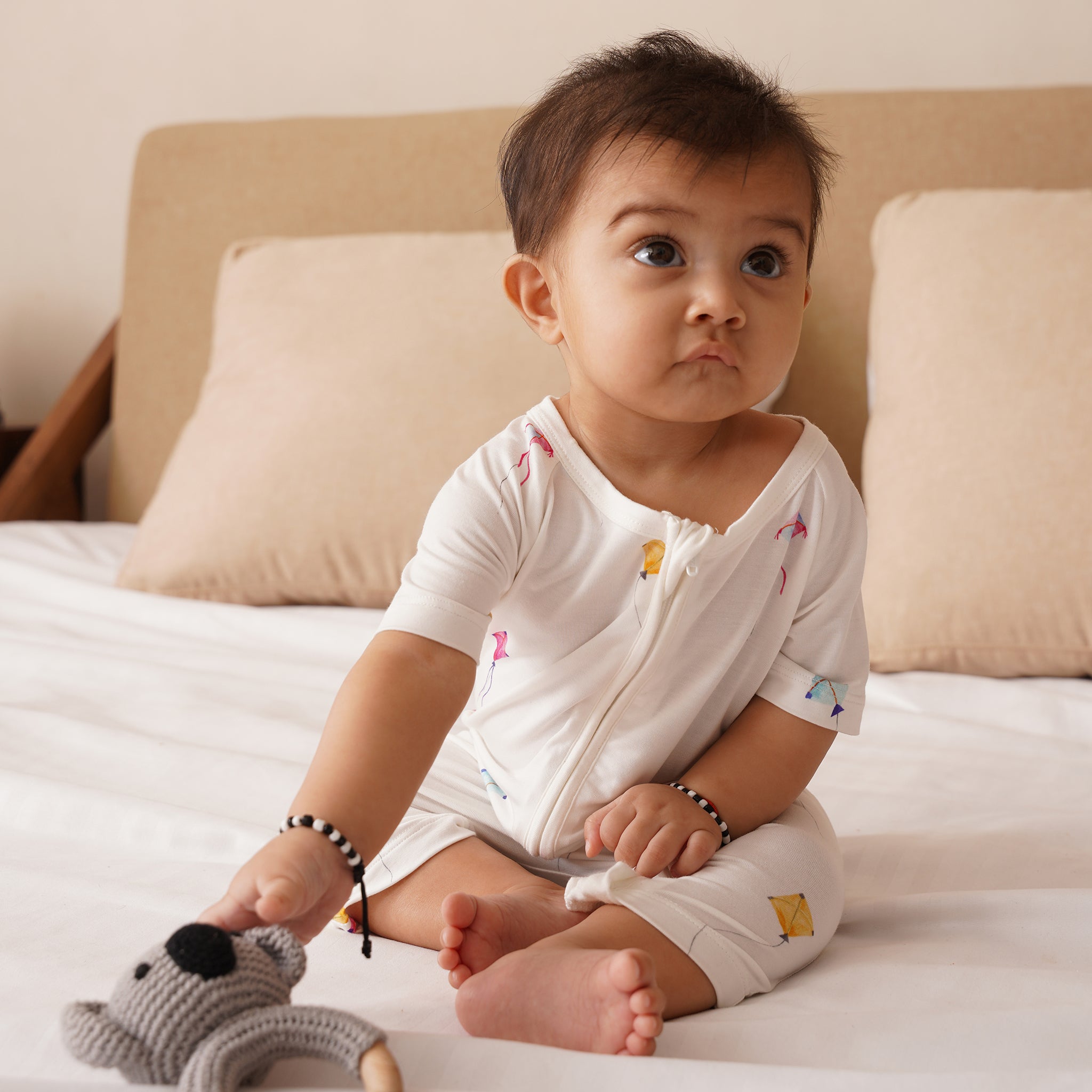 Baby sitting on a bed holding a toy, wearing a white bamboo outfit with colorful patterns.