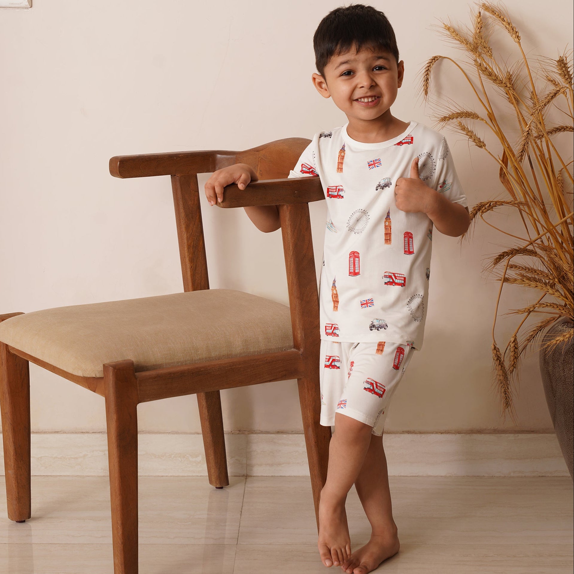 Child in a bamboo white shirt with red patterns standing next to a wooden chair indoors.