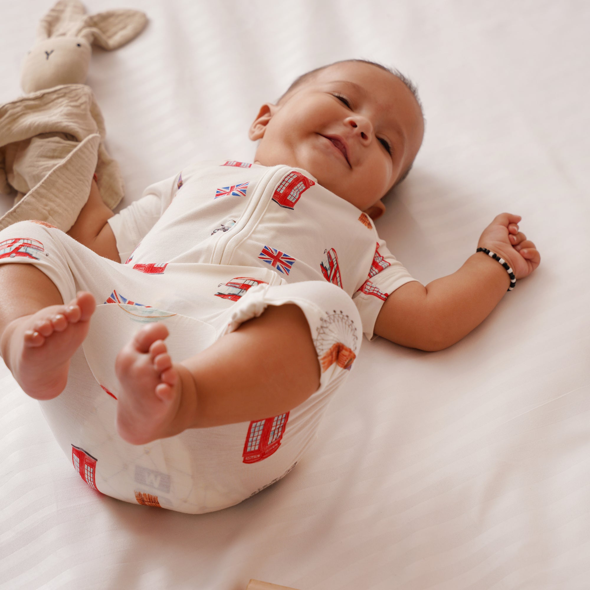 Baby lying on a white surface wearing a bamboo onesie with British flag pattern, next to a soft toy.