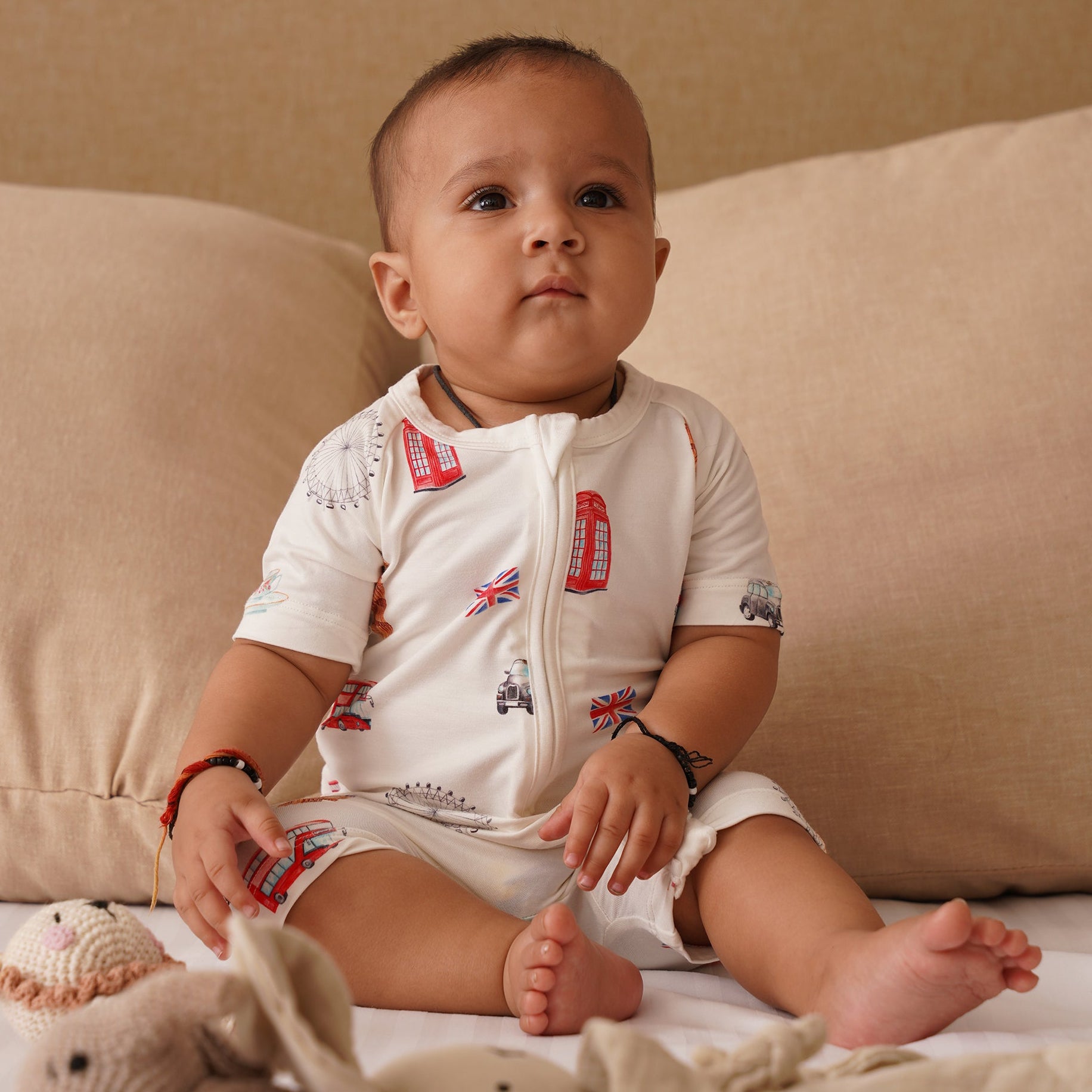 Baby sitting on a couch with toys around, wearing a white bamboo onesie with red and blue designs.