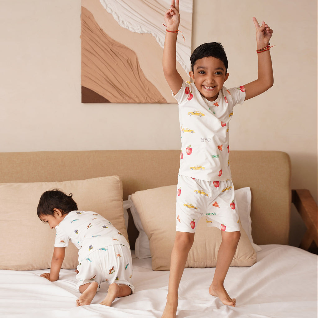 Two children in matching pajamas on a bed with a neutral-colored wall and artwork in the background.