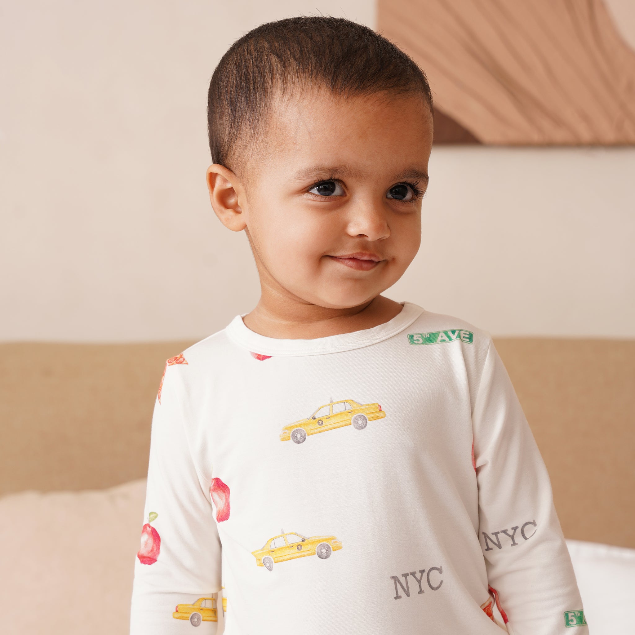 Child wearing a bamboo long-sleeve shirt with taxi and 'NYC' print, sitting on a bed.