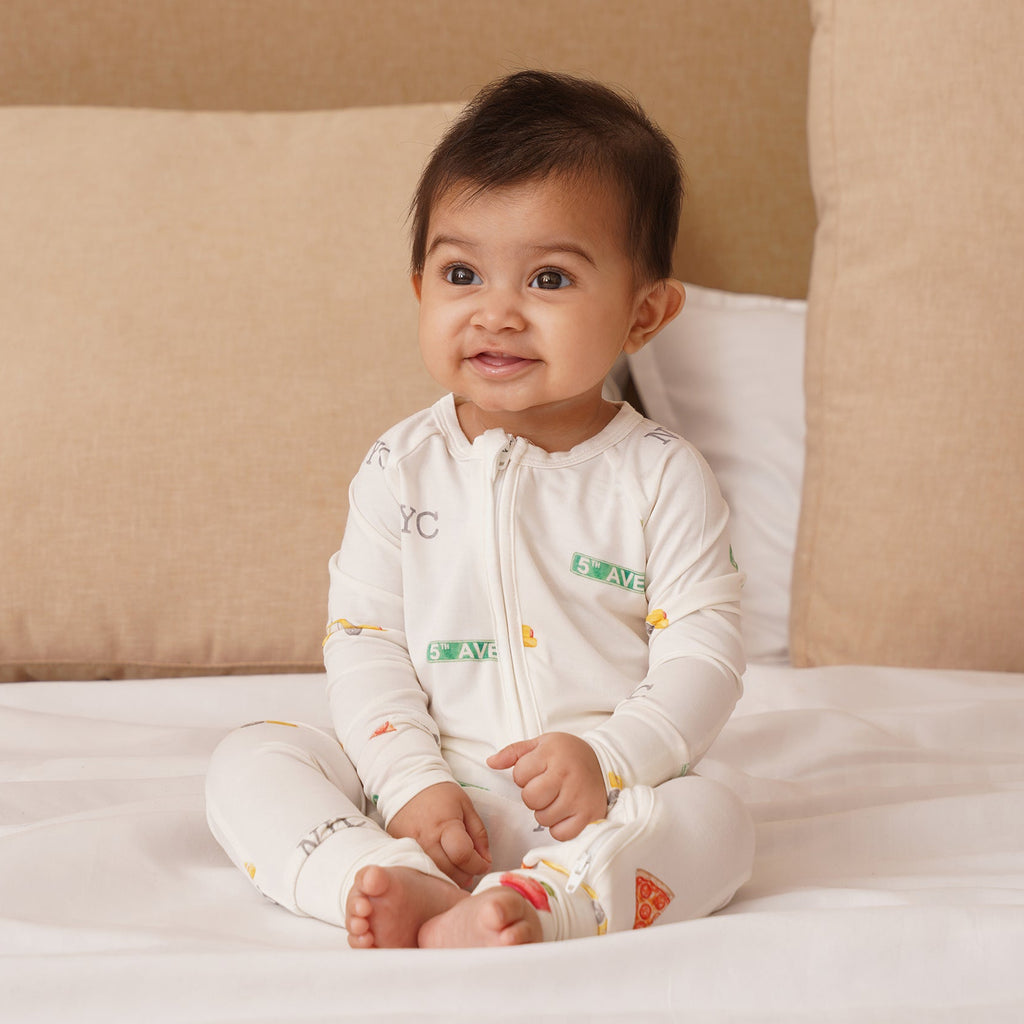 Baby sitting on a bed wearing a white bamboo onesie with colorful patterns.