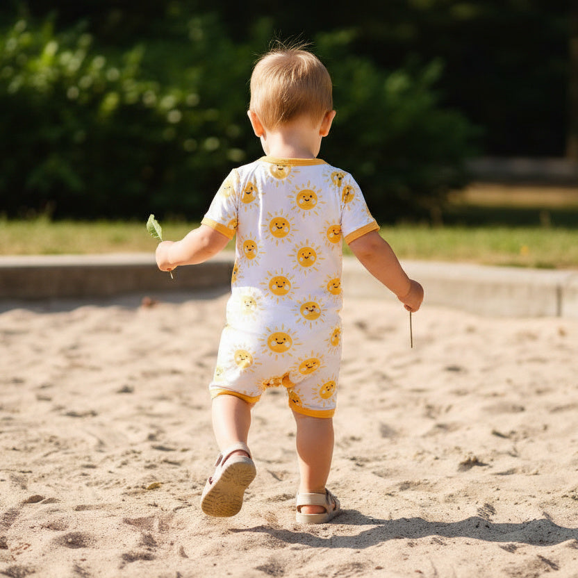 Child in a yellow and white romper walking on sand with greenery in the background