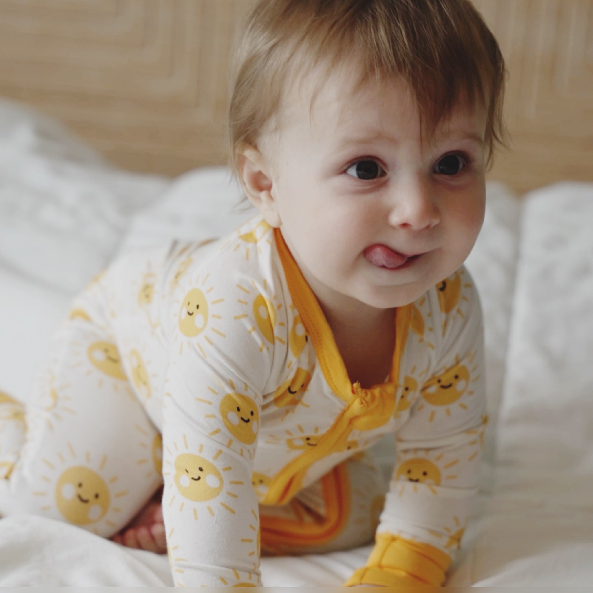 Baby wearing a yellow and white outfit with sun patterns on a bed.