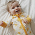 Baby wearing a yellow and white outfit with a zipper, lying on a white blanket.