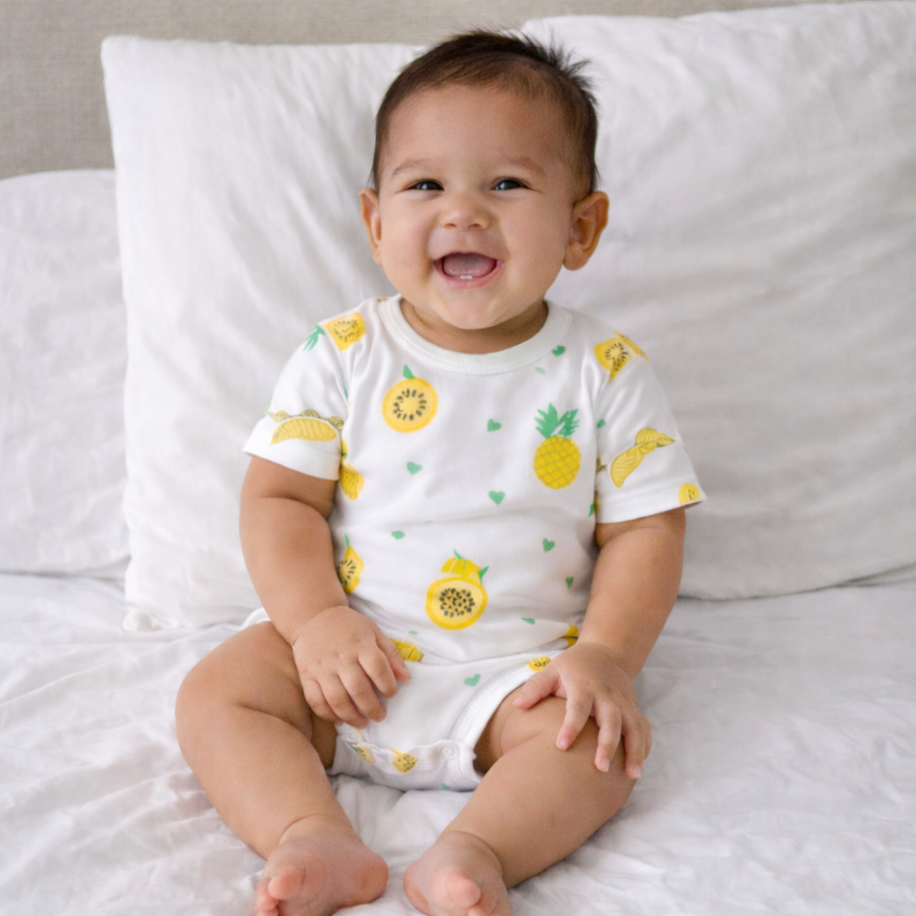 Baby wearing a white onesie with yellow fruit patterns, sitting on a white bed.