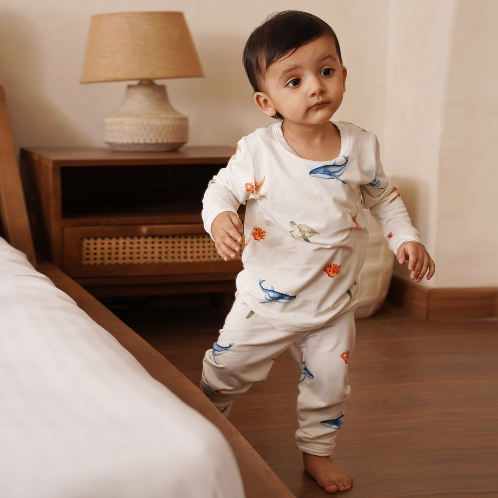 Baby in a white bamboo outfit with colorful designs standing in a bedroom.