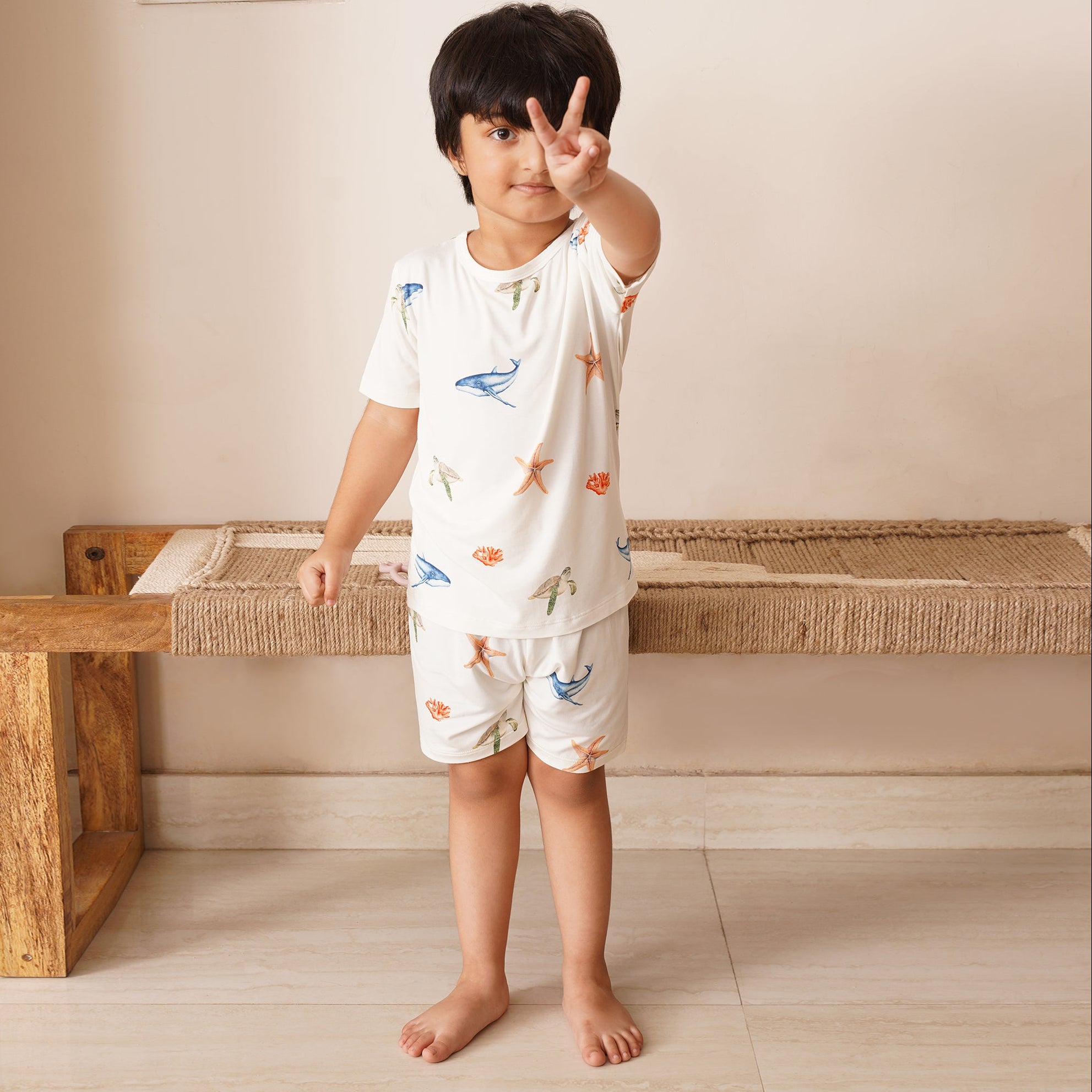 Child wearing a white  bamboo outfit with colorful animal prints, standing in a room with a wooden bench.