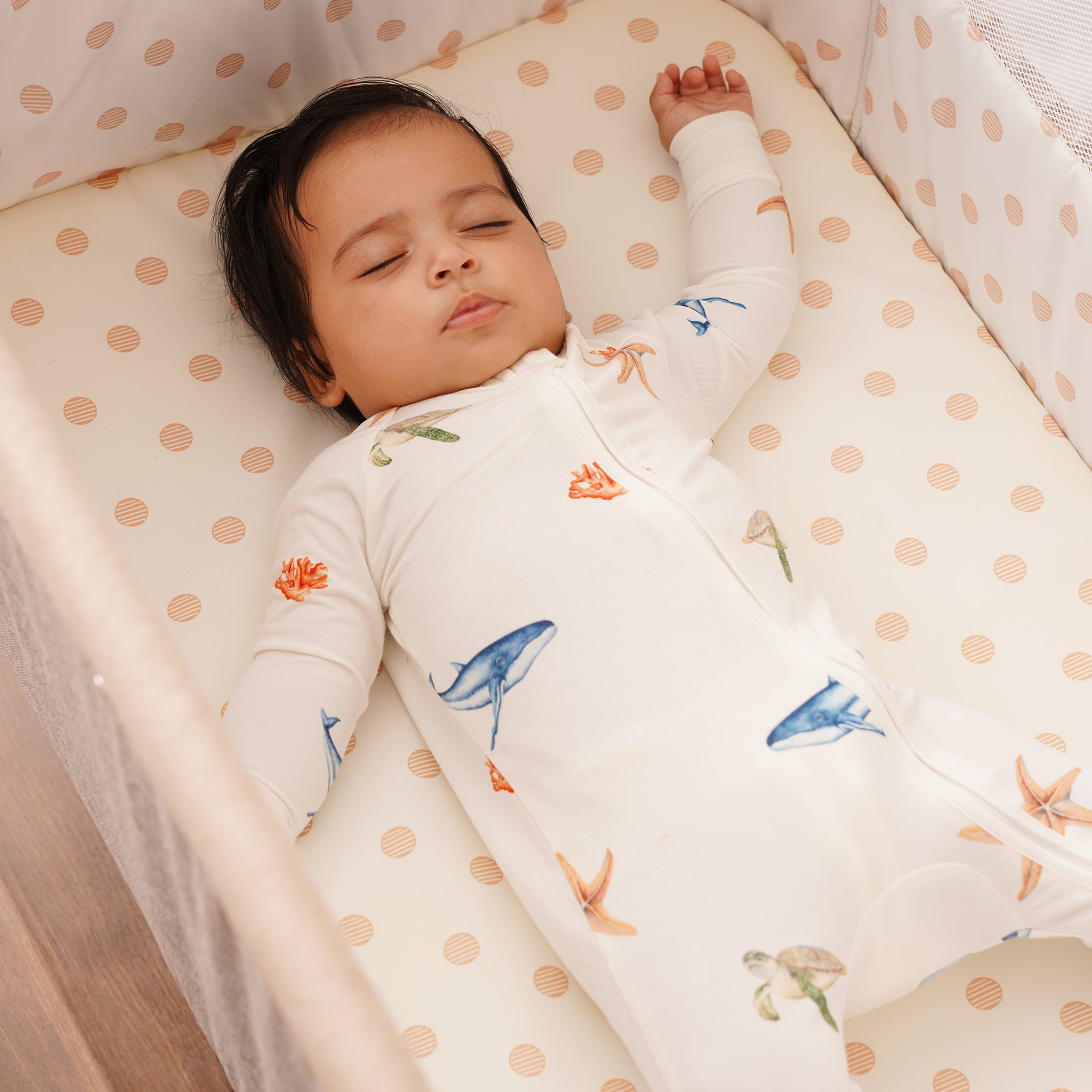 Baby sleeping in a crib with a polka dot sheet and a patterned blanket, wearing bamboo onesie