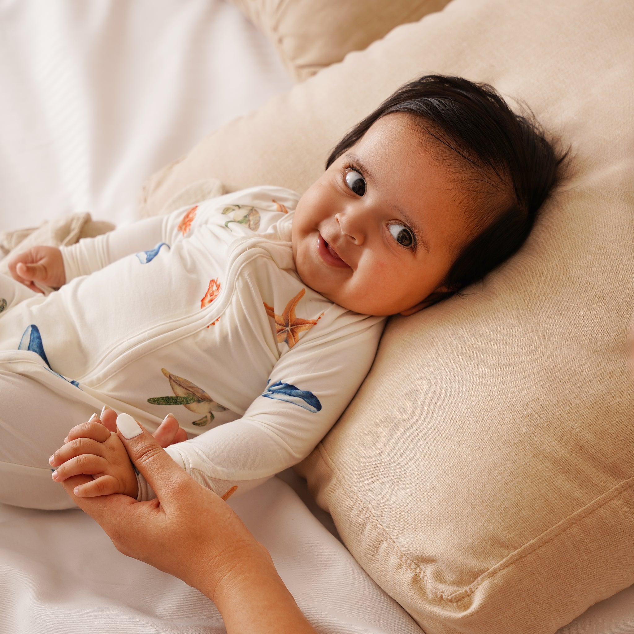 Baby lying on a beige pillow with a person holding their hand, wearing a white bamboo onesie with colorful patterns.